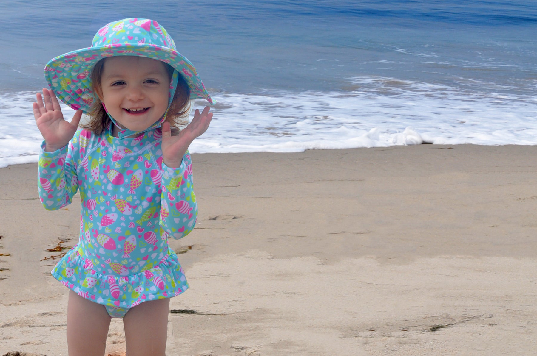 Young girl playing at the beach wearing the Flap Happy rainbow fish swimsuit and matching Flap Happy hat. 