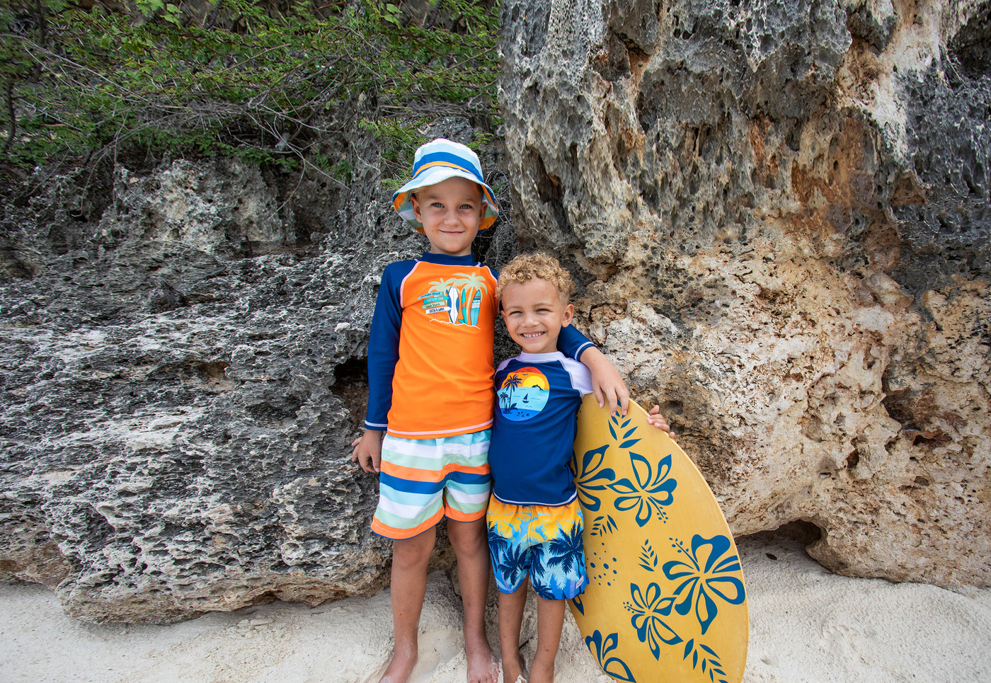 little brother and big brother on the beach wearing flap happy swim wear and hat holding surfbaord at the beach