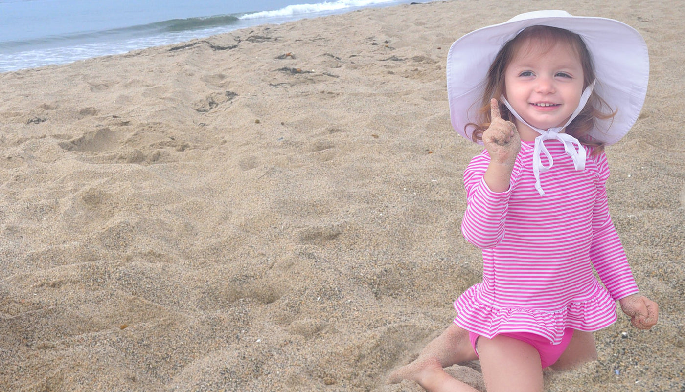 Little girl at beach wearing a white floppy sun hat and a pink and white stripe ruffle rash guard