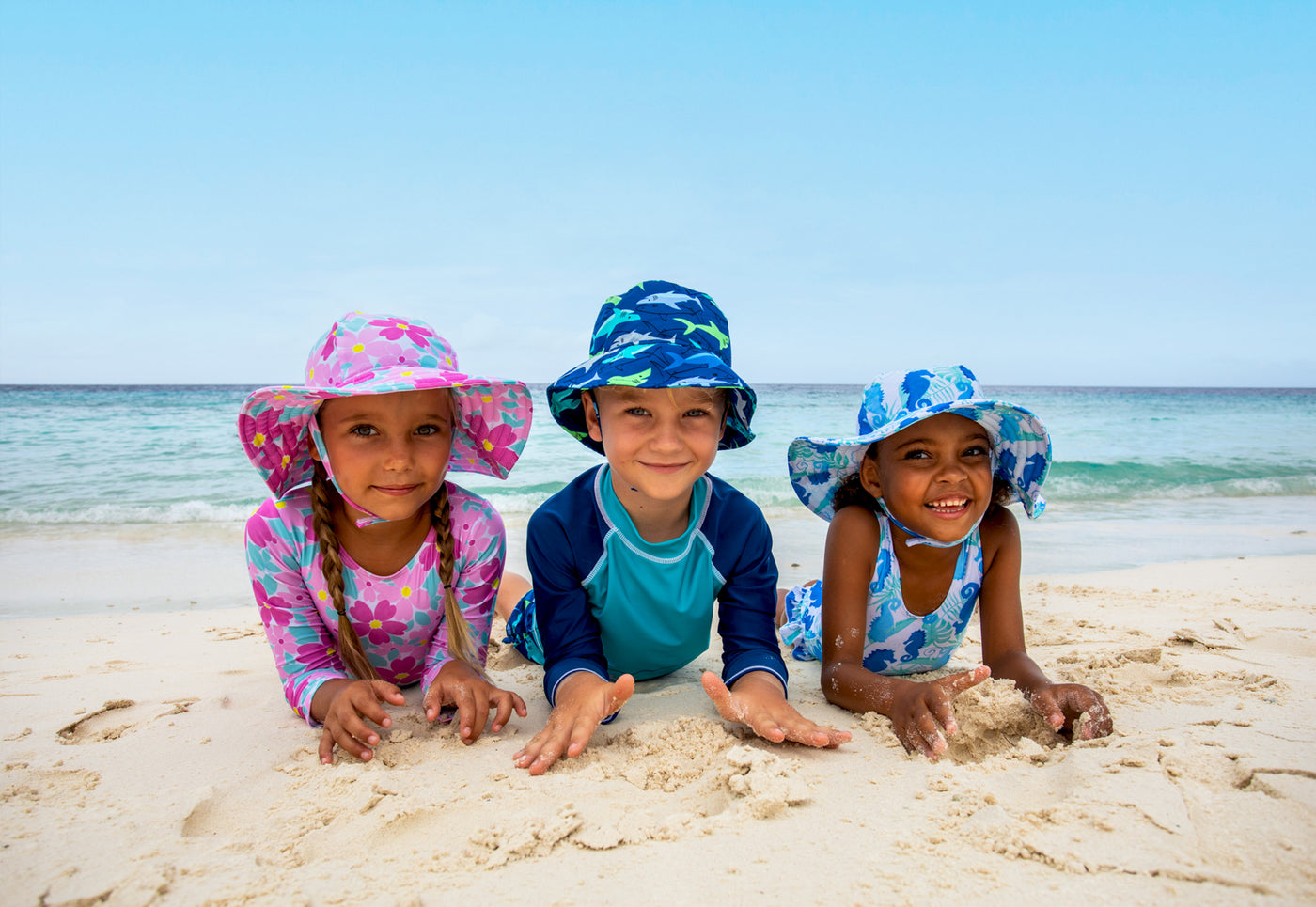 girl wearing swim hat and swimsuit in florals, boy wearing sun hat and swim in sharky and another girl wearing blue seashorse swim hat and swimsuit laying down on the beach