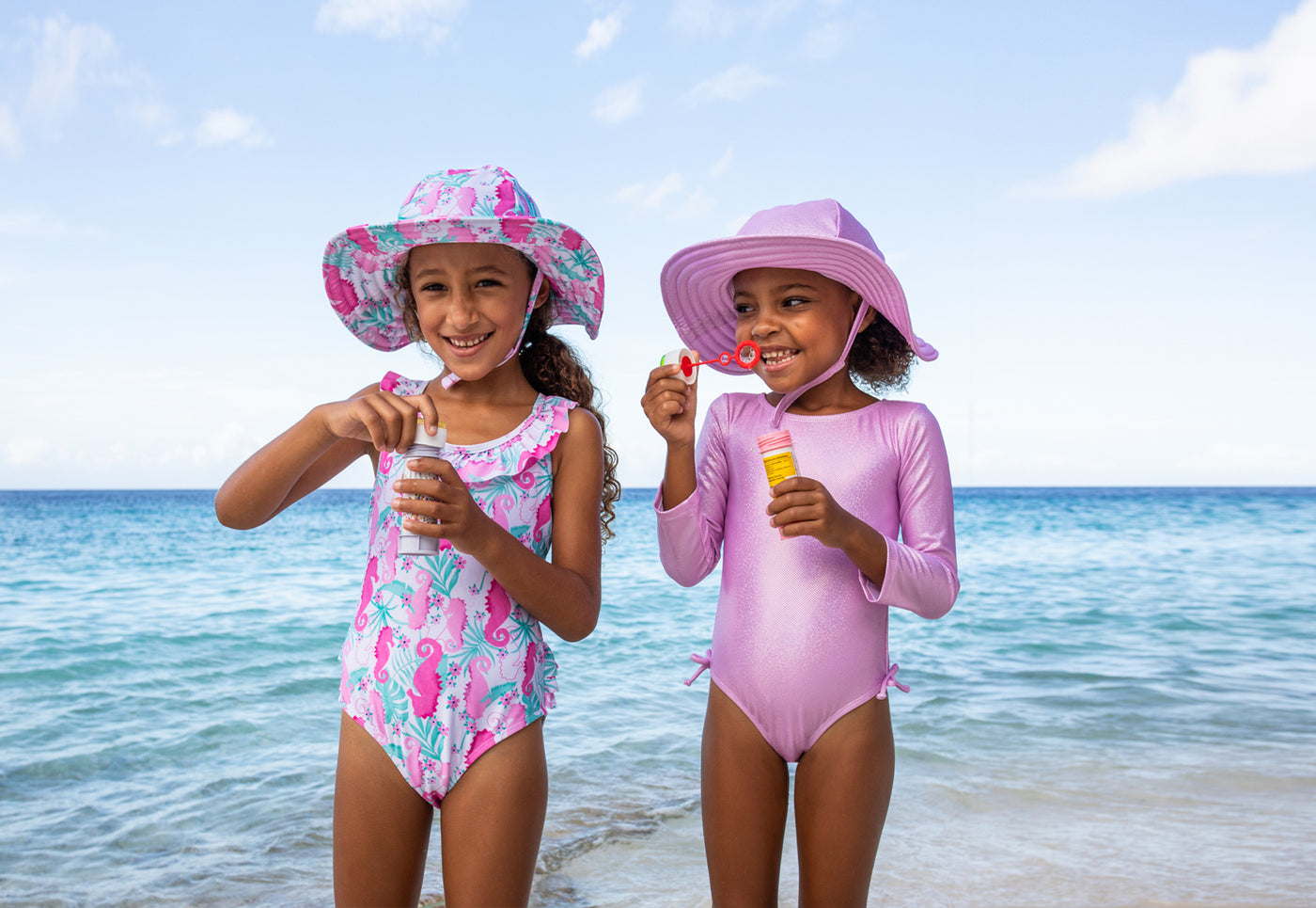 two girls wearing Flap Happy swimwear and hats blowing bubbles at the beach