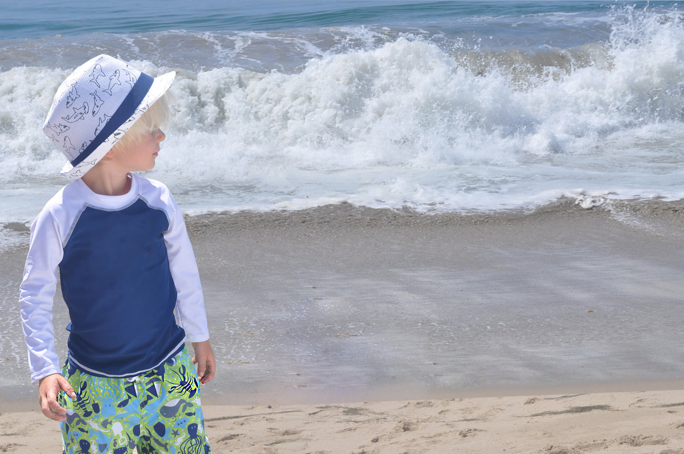 Young boy is standing at the beach in front of the ocean looking off to the right. Boy wearing matching white shark Flap Happy hat, blue and white long sleeve rash guard and green and blue crab print swim shorts.