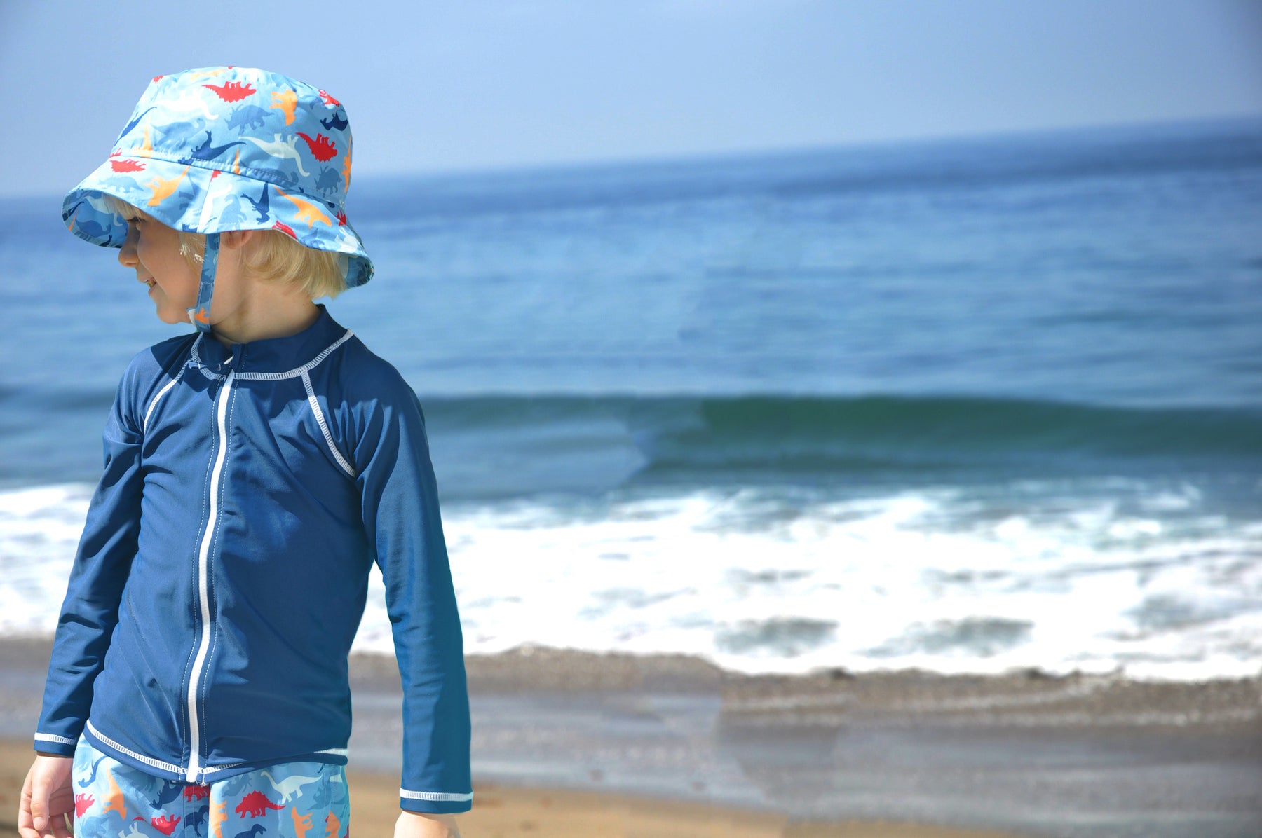 Young boy is standing at the beach in front of the ocean looking off to the left. Boy wearing matching blue dinosaur print Flap Happy bucket hat and shorts with blue front zip rash guard.