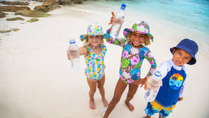 3 kids standing in the sand holding up water bottles. Girl 1 wearing pineapple print bathing suit and hat. Girl 2 is wearing a leopard and flower print bathing suit and hat. Boy on right wearing matching blue island bucket hat, shirt and shorts.